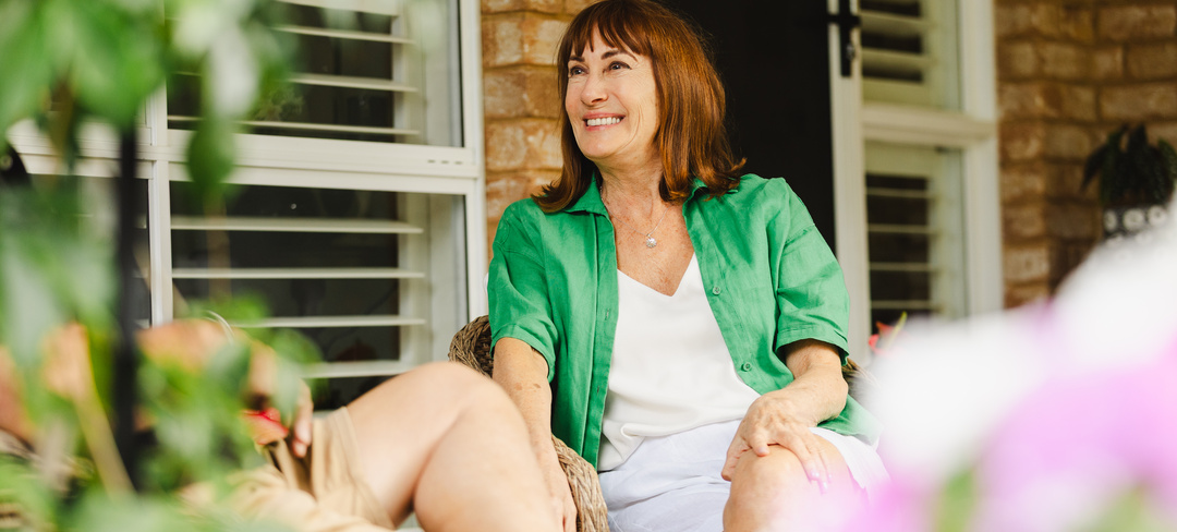 A female wearing a green blouse is sitting in front of a home smiling and chatting to a male 