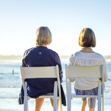 Two women sit on white folding chairs with their backs to the camera, looking out across a coastal view with swimmers in the ocean and a long beach in the distance across the water.