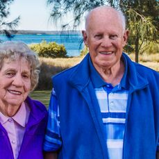 Retirement village residents Bruce and Dorothy Murray stand with beautiful Bonnells Bay in the background. Dorothy wears purple, and John wears bright blue. They’re smiling as they look at the camera.