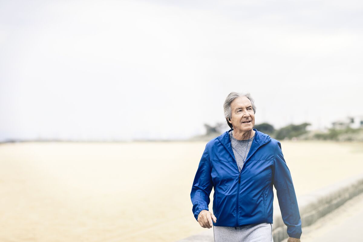 An older man in a blue jacket walking along a beach, listening to his headphones. 