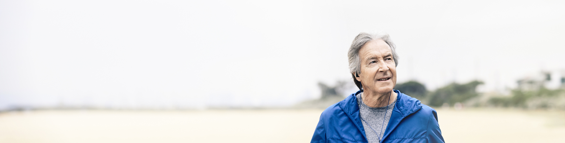An older man in a blue jacket walking along a beach, listening to his headphones. 