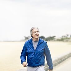 An older man in a blue jacket walking along a beach, listening to his headphones. 