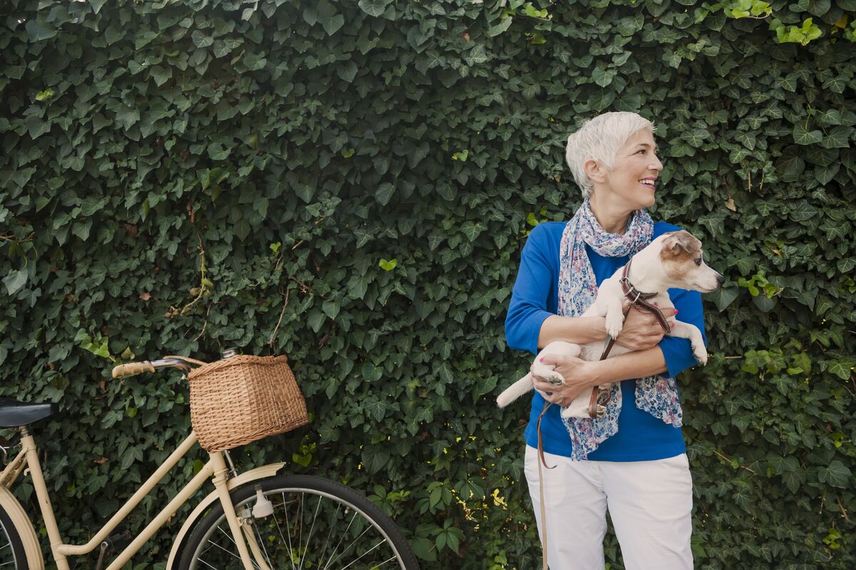 A woman holding a dog on her arm and standing in front of a hedge next to a bike