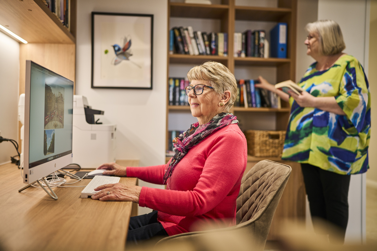 A senior lady sitting and using the computer while another lady is in the background surfing the bookshelf