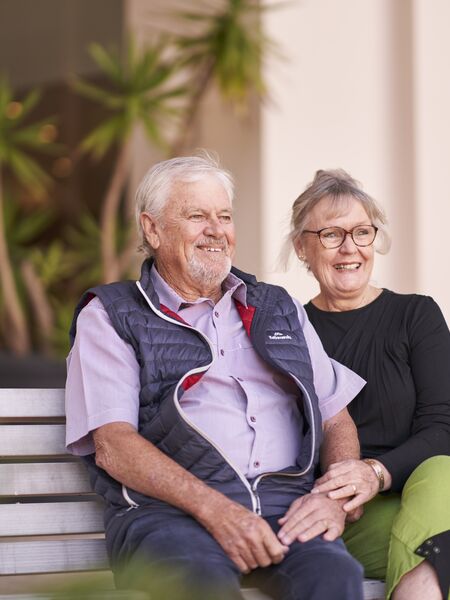 A male and female resident of Ardency Aroona at Yowie Bay sit on a bench in the garden and smile