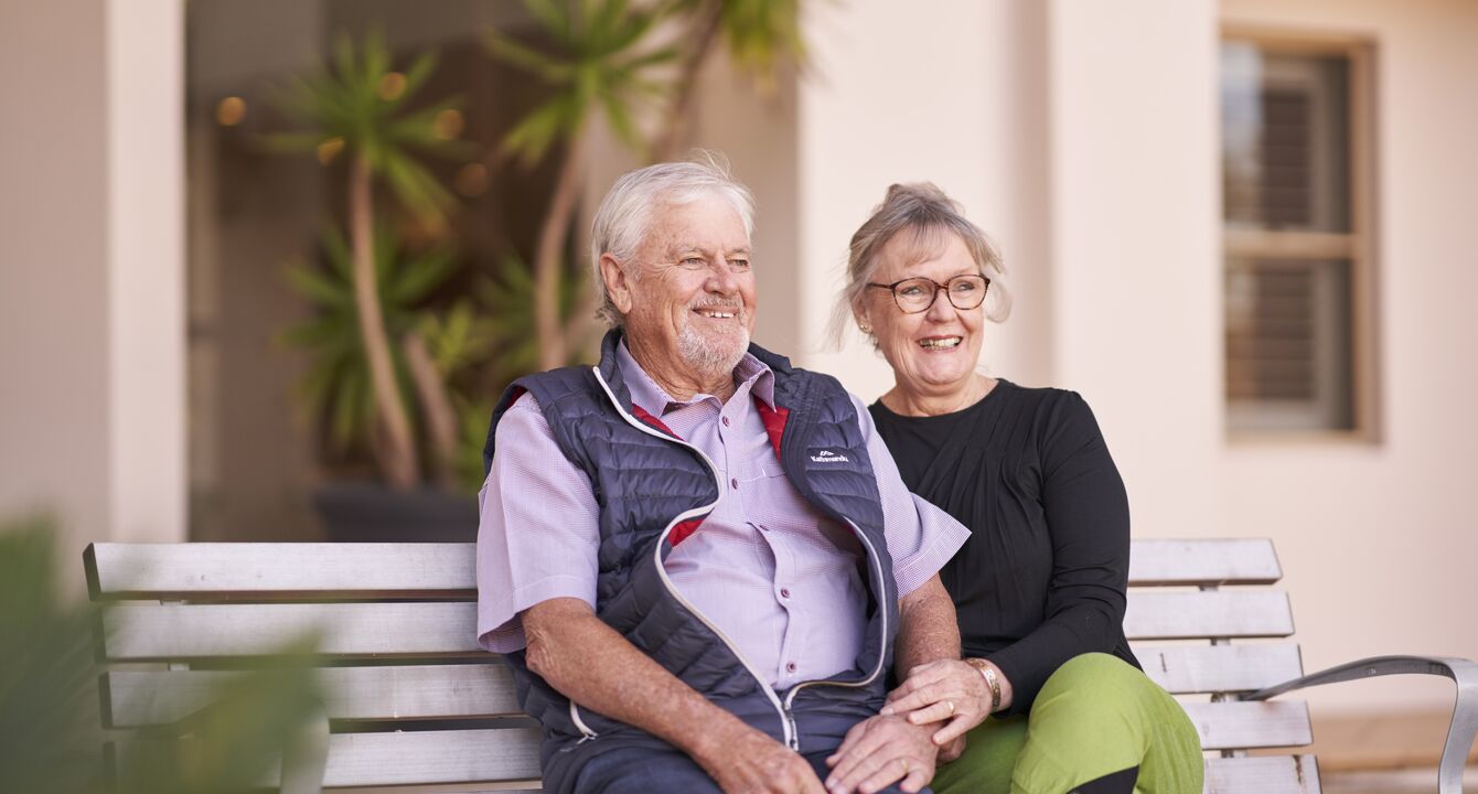 A male and female resident of Ardency Aroona at Yowie Bay sit on a bench in the garden and smile