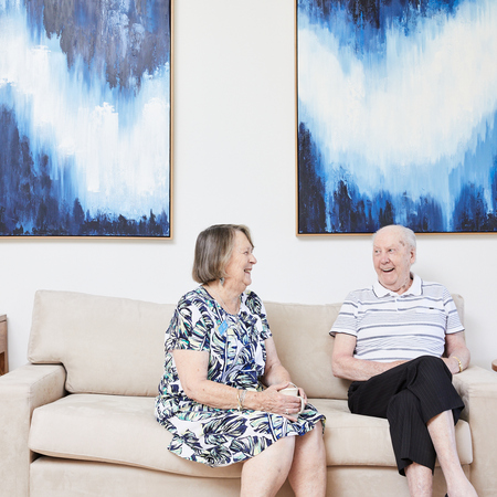 Two residents from Koorootang Court sitting on a couch in front of two blue artworks smiling at each other