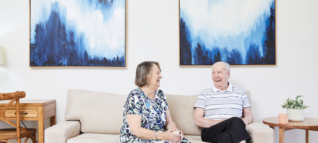 Two residents from Koorootang Court sitting on a couch in front of two blue artworks smiling at each other