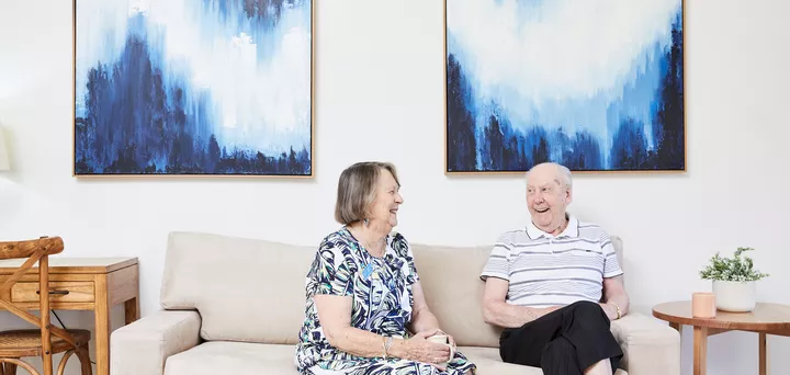 Two residents from Koorootang Court sitting on a couch in front of two blue artworks smiling at each other