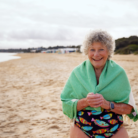 A retirement resident standing on a beach smiling wrapped in a green towel