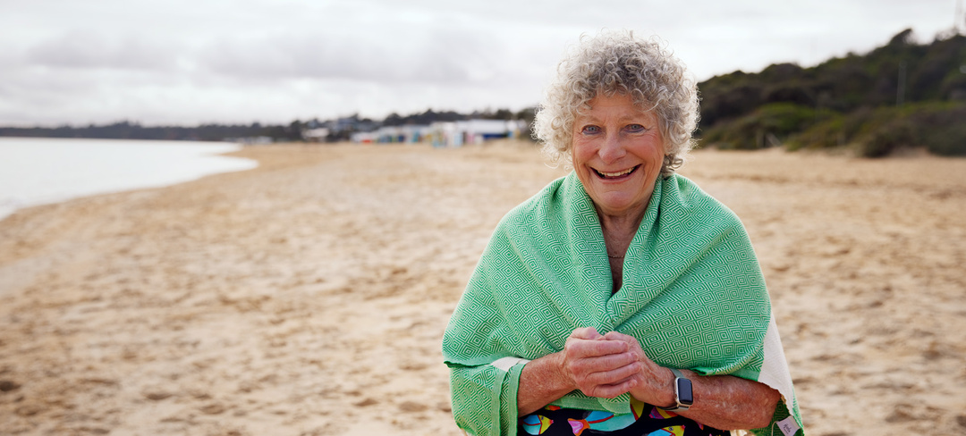 A retirement resident standing on a beach smiling wrapped in a green towel
