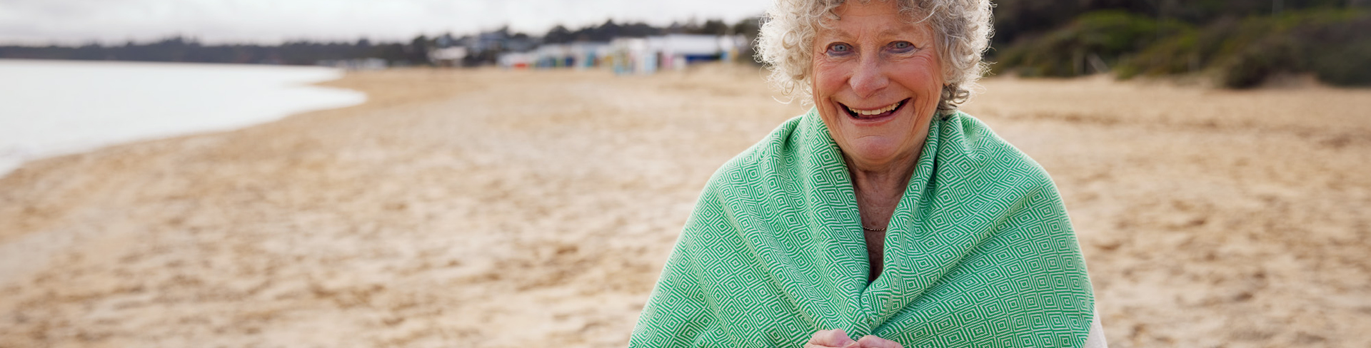 A retirement resident standing on a beach smiling wrapped in a green towel