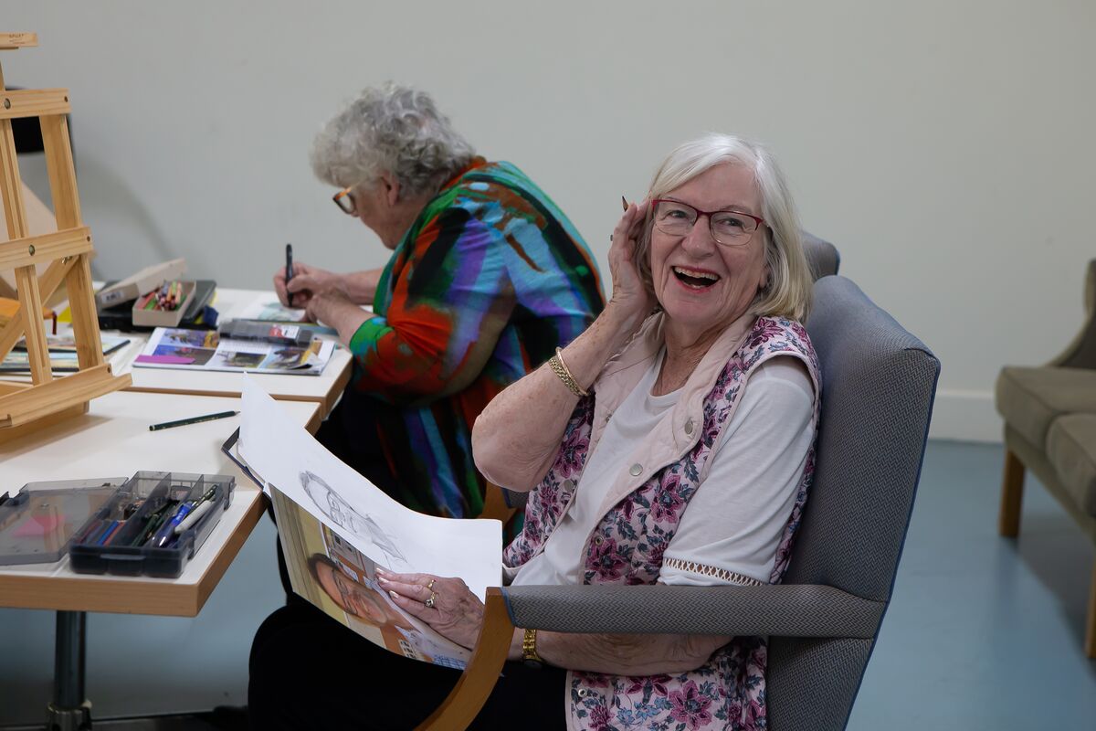 An elderly lady smiling while holding a drawn portrait in a retirement village arts room