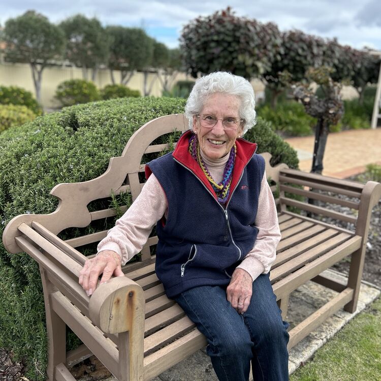 an old woman looking and smiling at the camera sitting on the bench outside