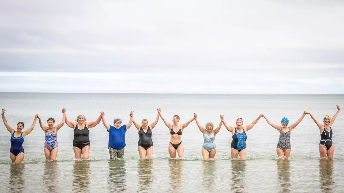 Members of the Sea Wolves stand in a line in knee-deep water. They form a chain holding each other’s hands aloft in a symbol of togetherness. There are women of all shapes, sizes and ages.