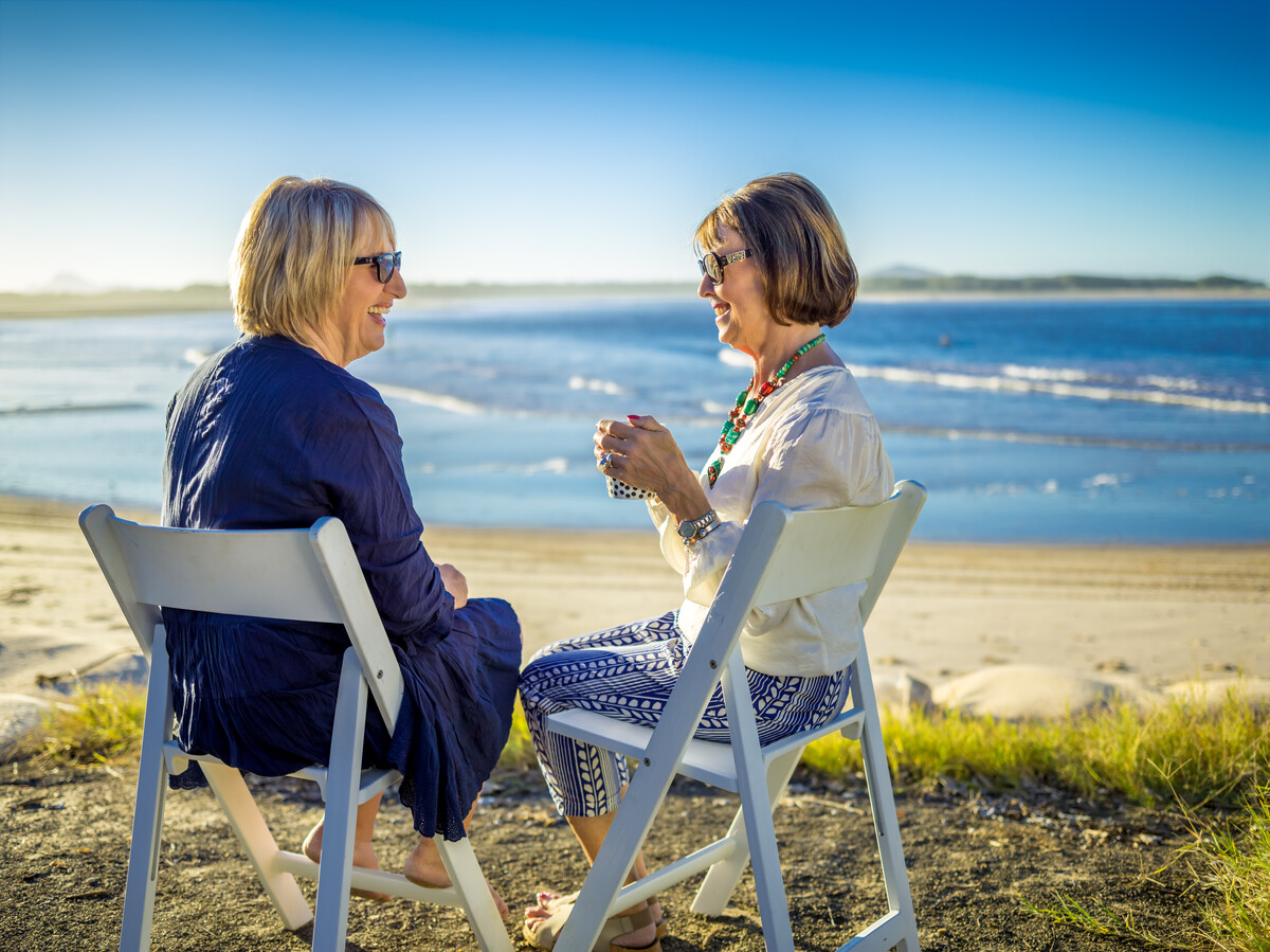 Two older women sitting in white chairs on the beach, smiling and engaging in conversation. 