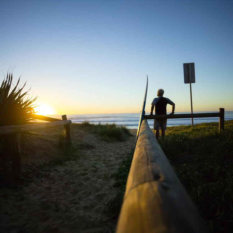 A man and his surfboard leaning on a fence, on the beach at sunset