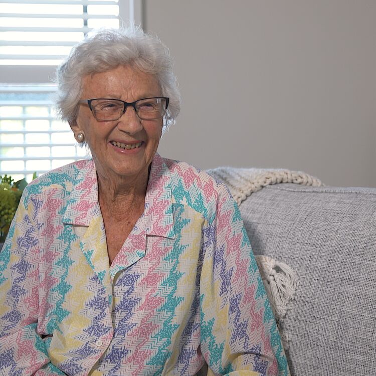 Resident June Neal sits on a grey sofa wearing a colourful blouse and smiling brightly.