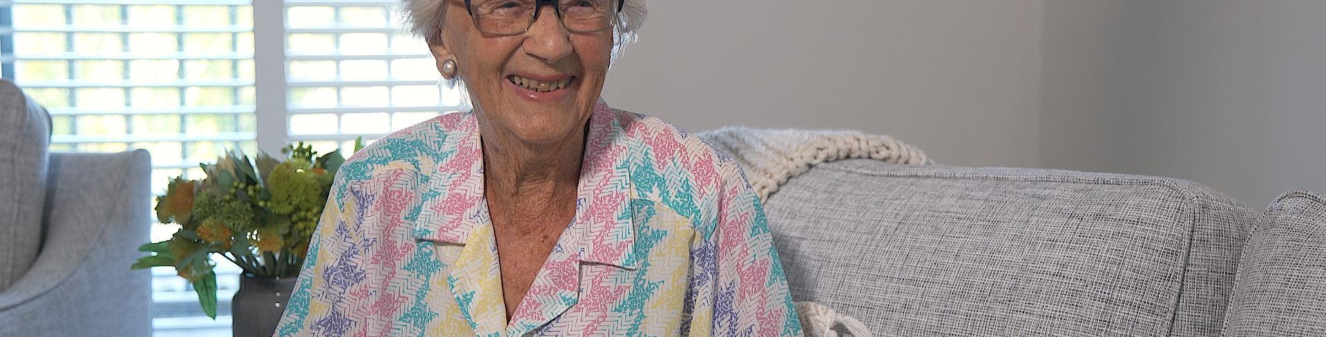 Resident June Neal sits on a grey sofa wearing a colourful blouse and smiling brightly.