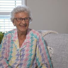 Resident June Neal sits on a grey sofa wearing a colourful blouse and smiling brightly.