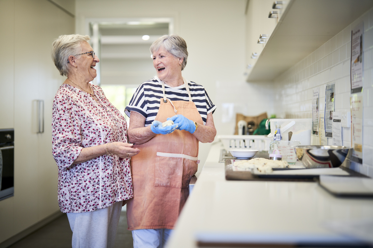 Two female retirement village residents standing in the kitchen forming dough and laughing