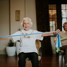 An older lady sitting on a chair and doing arm exercises with an elastic band