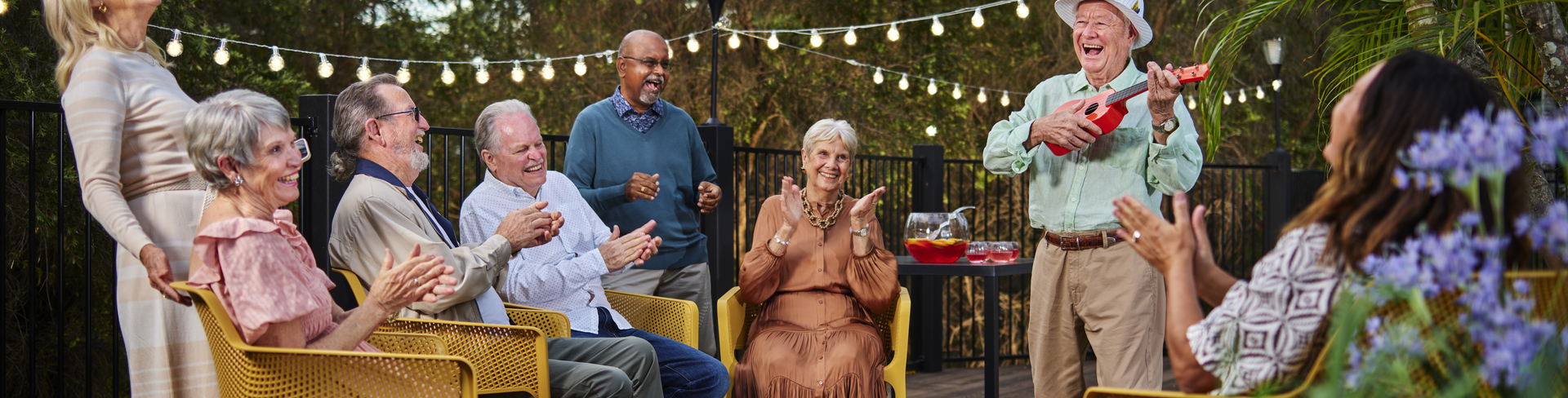 A group of retirement village residents clapping for an elderly man playing the ukulele.