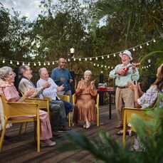 A group of retirement village residents clapping for an elderly man playing the ukulele.