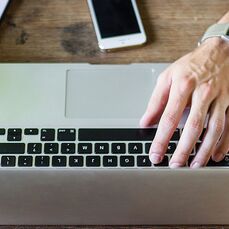 A top view of a person’s hands using a laptop open on a desk, with one hand typing and the other writing on a pad. 