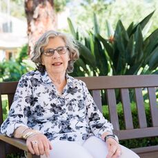 Retirement village resident Robin sits on a park bench wearing a black and white floral blouse. She has wavy grey hair and wears glasses. 