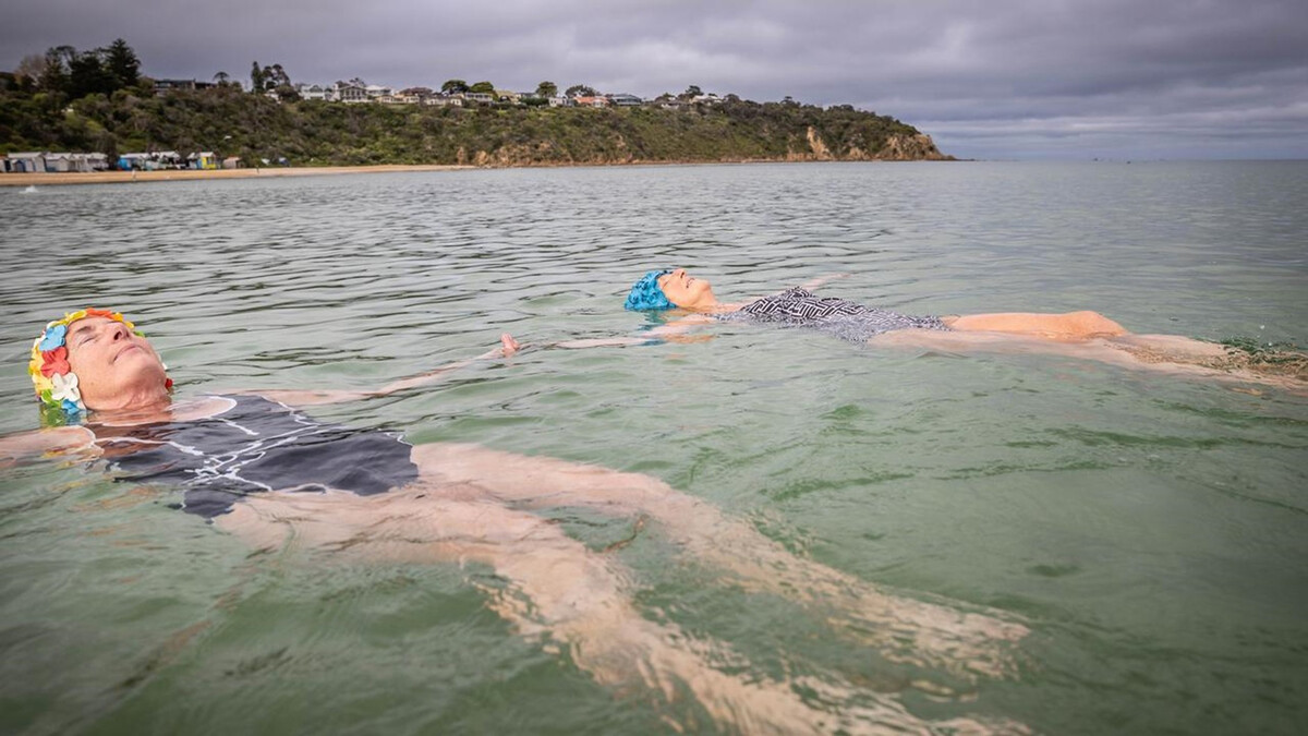 Two women float on their backs in the cold waters of Port Phillip Bay. They’re wearing bathing suits and colourful floral swimming caps.