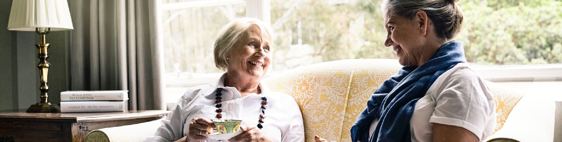 Two women sit on a yellow and white lace-patterned couch and looking at each other, enjoying a cup of tea and a chat. 