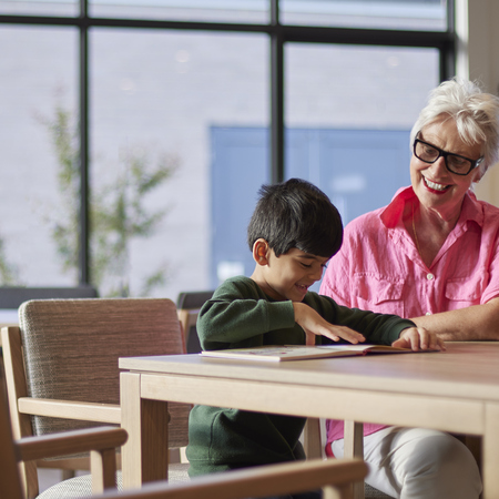 An elderly lady with glasses sits at a table with a young boy reading a book