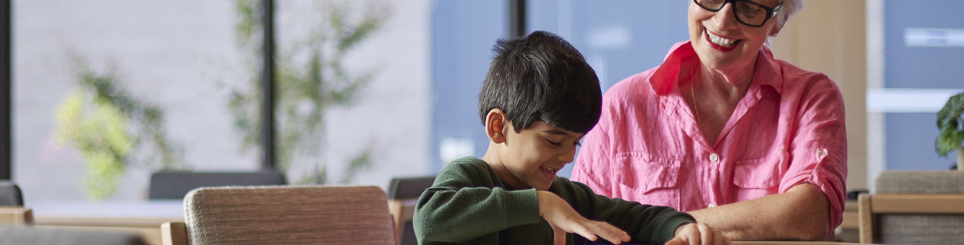 An elderly lady with glasses sits at a table with a young boy reading a book