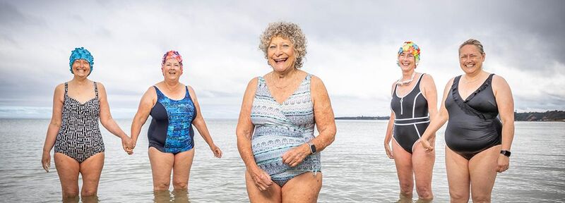 Five women stand in knee-deep water, all wearing bathing suits. Some wear colourful floral swimming caps. They’re smiling and holding hands.