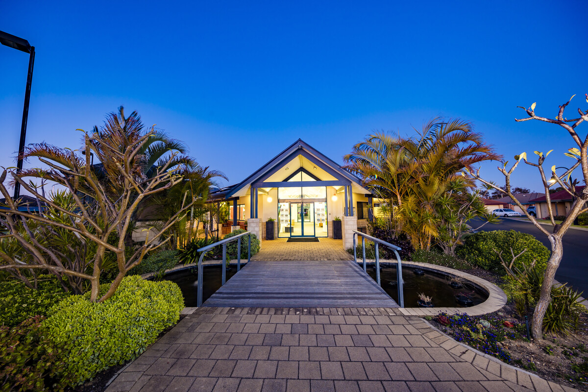 Woodstock West evening image of well lit main entrance outside with landscaped garden, walkway and walking bridge