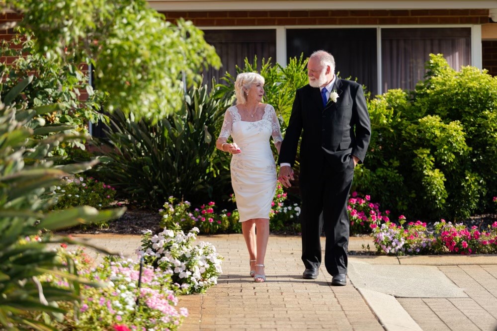 A senior couple, dressed in their wedding attire, stroll hand-in-hand down an outdoor path in a retirement village, smiling at each other.
