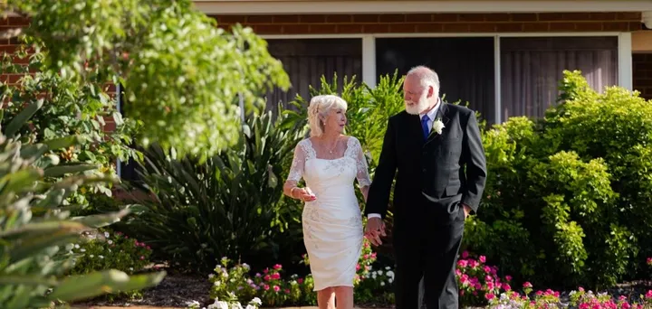 A senior couple, dressed in their wedding attire, stroll hand-in-hand down an outdoor path in a retirement village, smiling at each other.