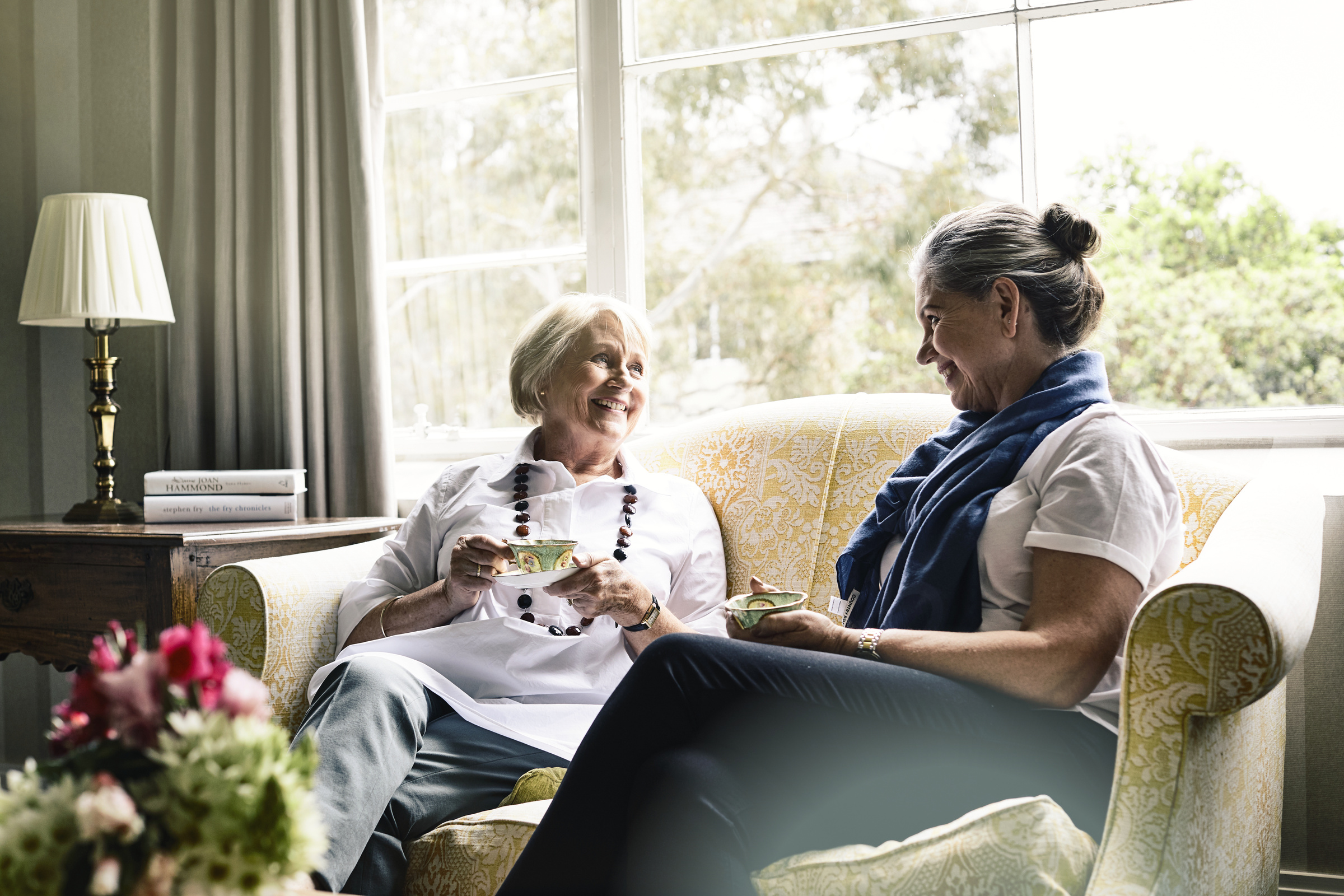 Couple of senior ladies having a cup of tea on the couch and smiling at each other