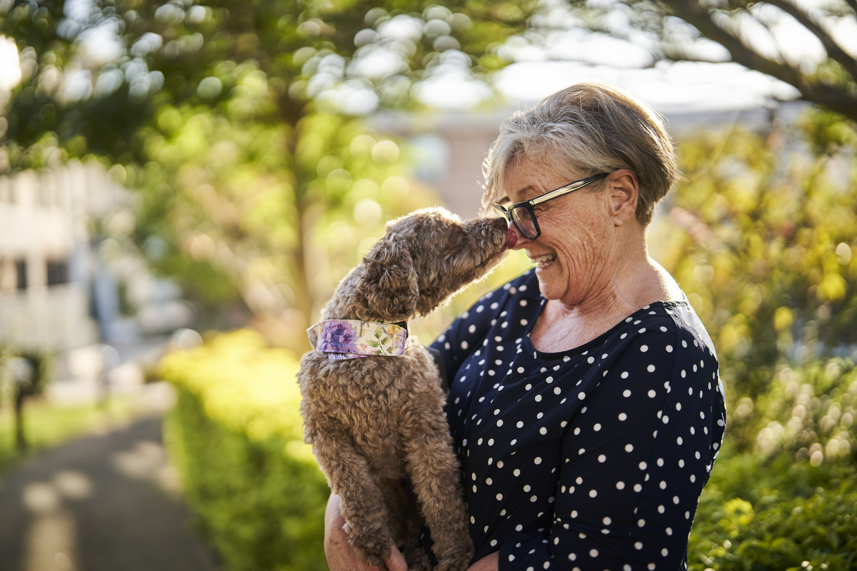 A women smiling and holding a small dog on her arm that is licking her nose