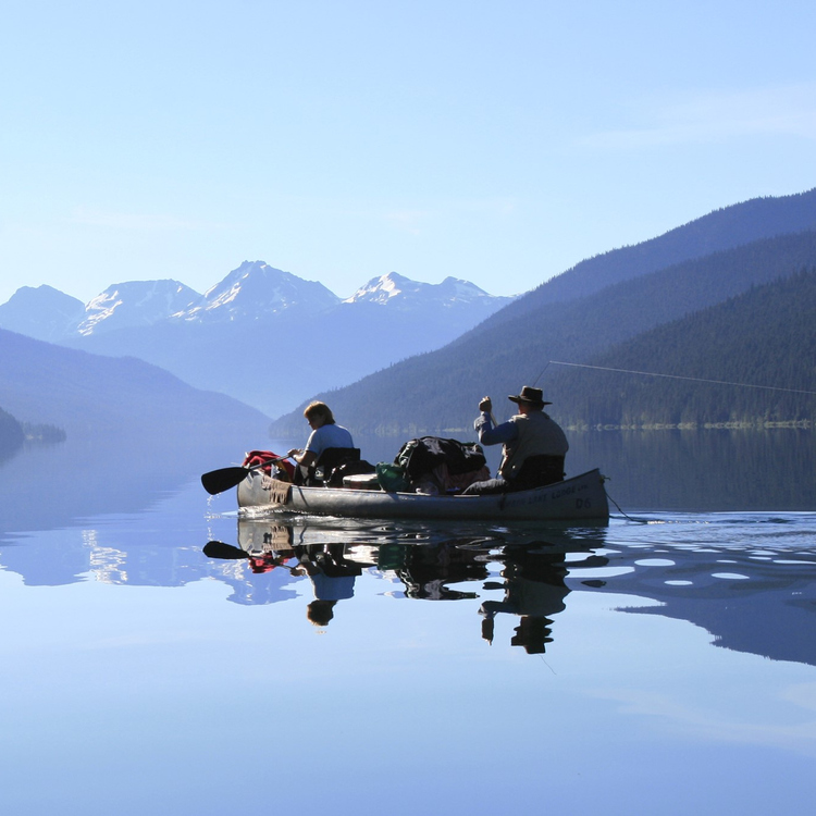 A photo of a crystal clear lake, where a few people are rowing a boat. The still water mirrors the expansive sky above and the mountains around the lake.