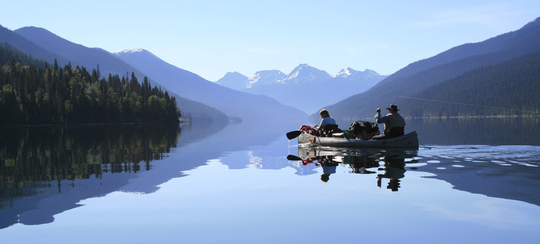 A photo of a crystal clear lake, where a few people are rowing a boat. The still water mirrors the expansive sky above and the mountains around the lake.