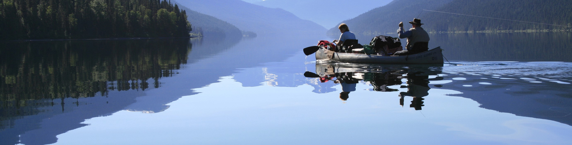 A photo of a crystal clear lake, where a few people are rowing a boat. The still water mirrors the expansive sky above and the mountains around the lake.