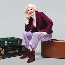 A smiling elderly woman sitting on a tan suitcase and wearing a striking maroon jacket and matching lipstick, looking ready to set off on her solo travels.