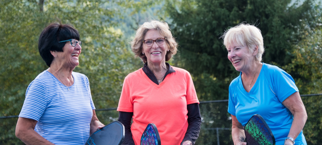 Three elderly ladies standing on a Pickleball court and holding Pickleball rackets