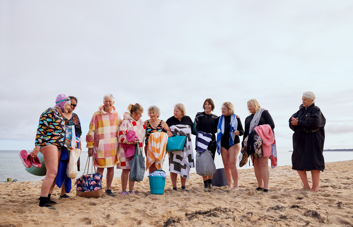 Five women stand in knee-deep water, all wearing bathing suits. Some wear colourful floral swimming caps. They’re smiling and holding hands.