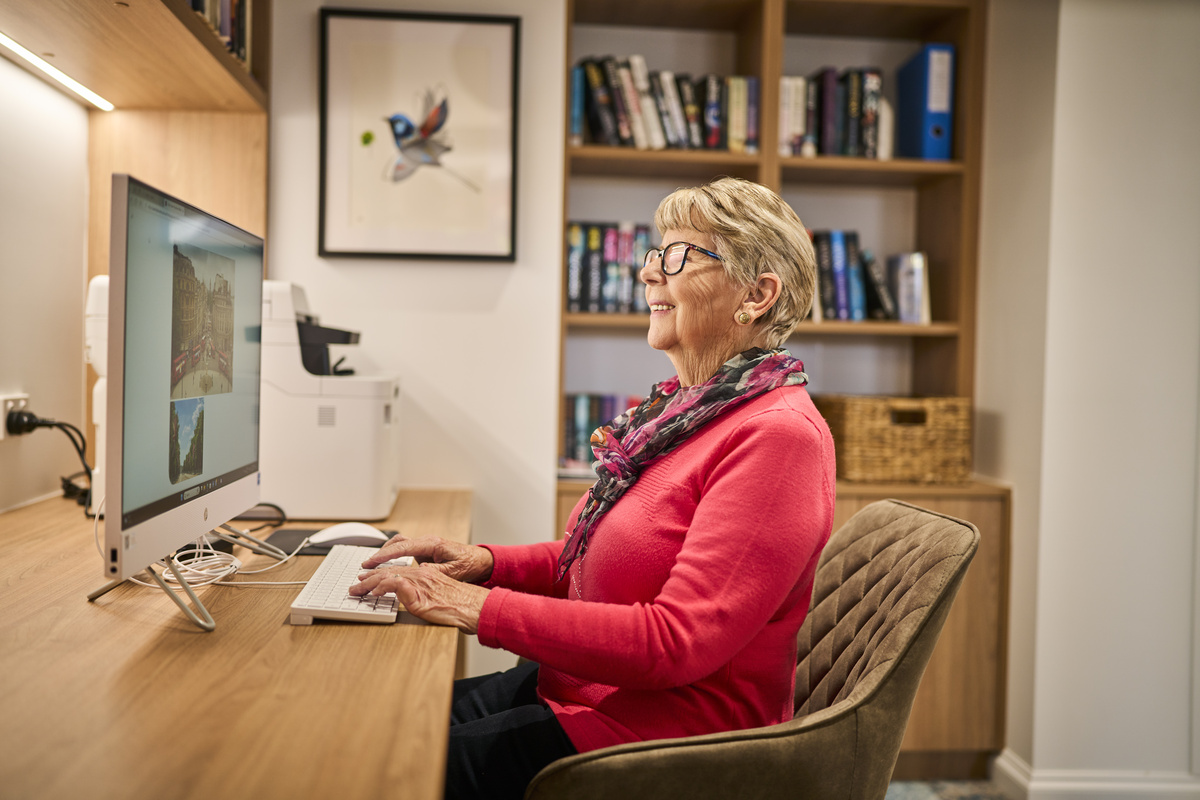 A lady in a red cardigan sitting on her computer in the study of her retirement apartment