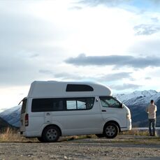 A camper van parked with a man standing nearby looking out across a lake at a view of snow capped mountains
