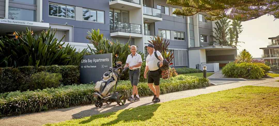 Two men walking through an apartment retirement village carrying golfing equipment