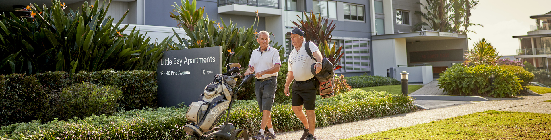 Two men walking through an apartment retirement village carrying golfing equipment